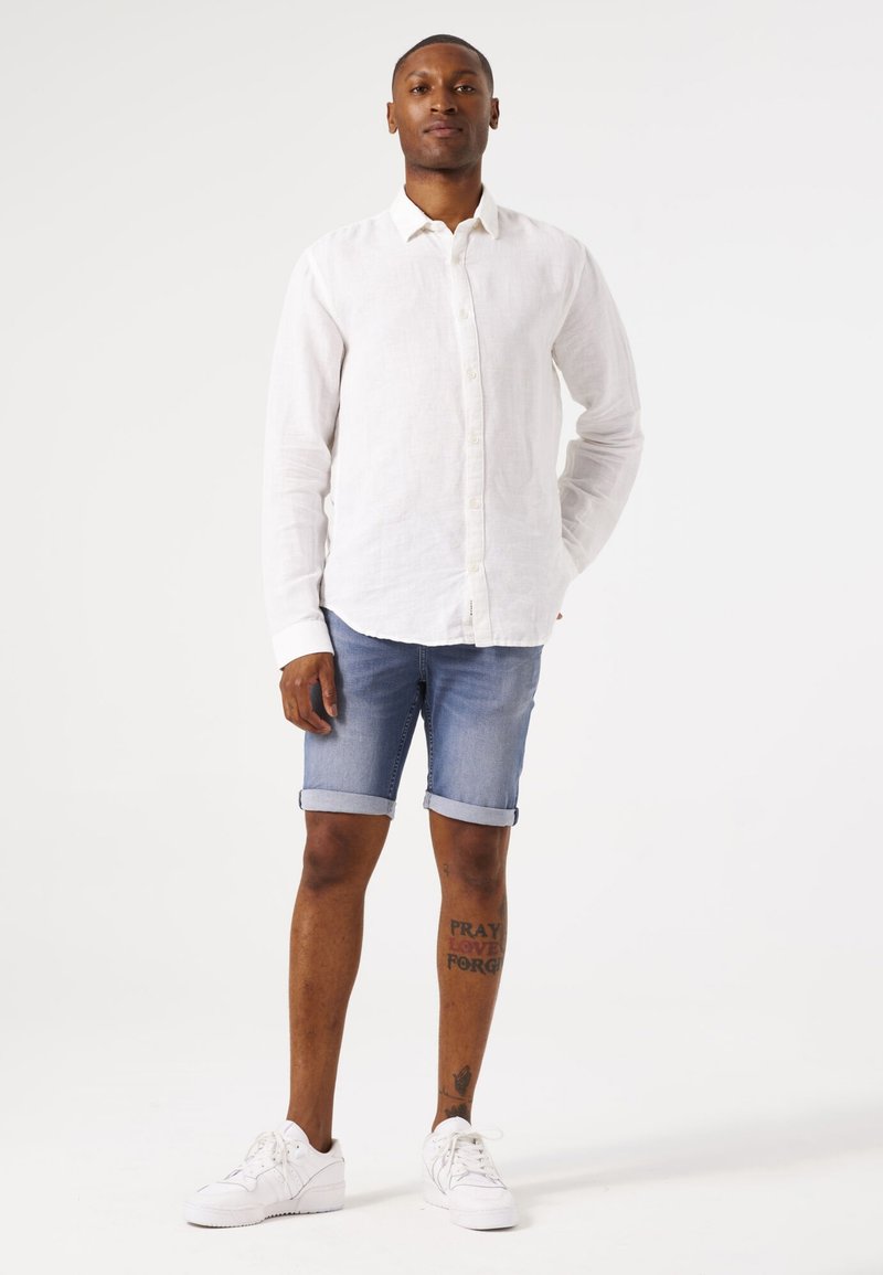 Chemise en lin blanche à boutons, shorts en denim avec ourlets roulés et baskets blanches ; tatouages visibles sur la jambe inférieure. Tenue simple et décontractée.