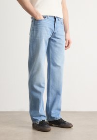 Ljust blå denimjeans med avslappnad passform, raka ben, synliga sömmar och fransade fållar, kompletterade med svarta sneakers.
