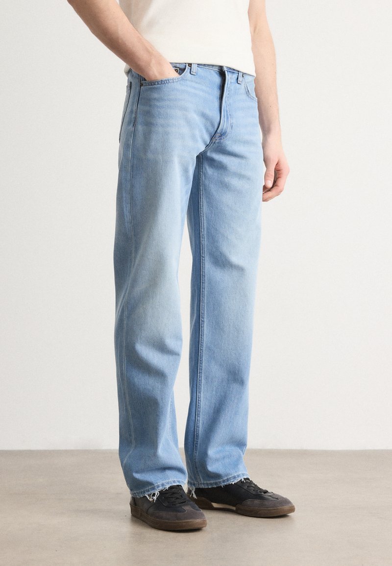 Ljust blå denimjeans med avslappnad passform, raka ben, synliga sömmar och fransade fållar, kompletterade med svarta sneakers.