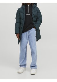 Cappotto imbottito verde con cappuccio, chiusura zip, indossato sopra un hoodie nero con lettering bianco, abbinato a jeans azzurri e sneakers bianche.