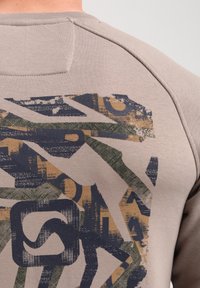 Beige sweatshirt featuring a textured, abstract pattern in blue, green, and gold on the back. Soft fabric with a round neckline and stitched details.