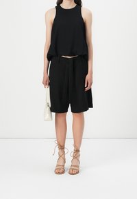 Mustat hihattomat lyhyet topit, väljät mustat shortsit solmimisvyöllä, beige nauhateräksiset sandaalit ja pieni valkoinen käsilaukku. Yksinkertainen tausta.