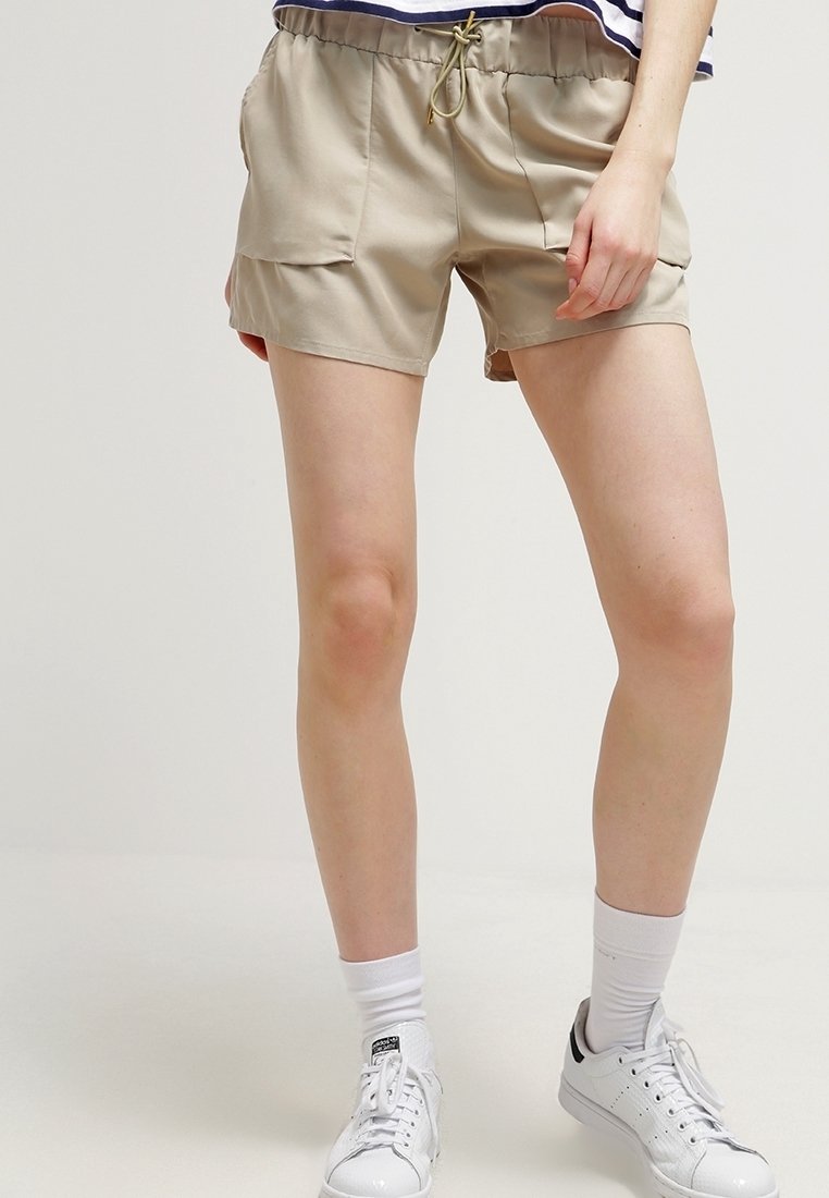 Beige shorts with an elastic waistband and drawstring. Features two front pockets, made of lightweight fabric. Worn with white sneakers.