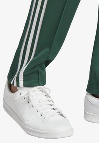 Green athletic pants with three white stripes along the side, paired with white leather sneakers featuring perforated detailing and lace closure.