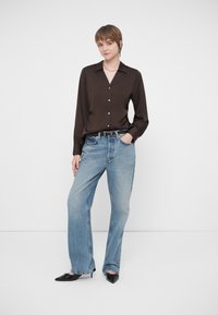 Noor inimene seisab, üks käsi taskus, seljas tumedast pruunist nööpidega särk, heledad sinised teksad, must vöö ja mustad teravad kingad.