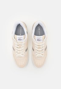 Beige joggesko med perforert design, hvite lisser, grå detaljer og teksturert overflate. Lacoste-logo på tungen og siden. Mykt materiale.