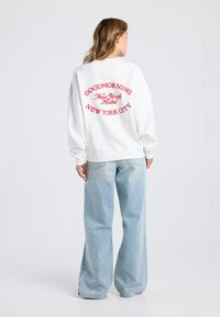 White sweatshirt with red embroidered text on the back reading "GOODMORNING New York Hotel" and "NEW YORK CITY"; paired with light blue wide-leg jeans.