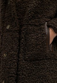 Manteau brun texturé en matériau duveteux, avec de grands boutons et une poche zippée bordée de cuir sur le côté.