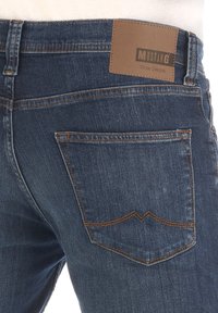 Vaqueros de mezclilla azul oscuro con una textura suave, que cuentan con costuras contrastantes en naranja y una etiqueta de cuero marrón con la inscripción "Mustang True Denim" en la parte de atrás.