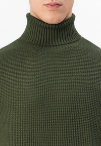 Nahaufnahme einer Person, die einen olivgrünen, gerippten Rollkragenpullover mit einem Waffelmuster trägt, mit Fokus auf den Hals und die obere Brust.
