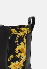 Acercamiento a una bota negra con paneles de cuero negro y una sección de tela que presenta cadenas barrocas doradas y patrones florales.