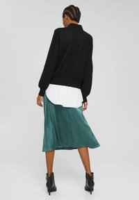 Vrouw met gevlochten haar in een zwarte trui, witte blouse, tealcoupe satijnen rok en zwarte enkellaarsjes die met de rug naar de camera staat op een grijze achtergrond.
