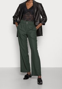 Pantalons larges verts avec une taille élastique, associés à un haut en dentelle noire et un blazer en cuir noir. Des mocassins noirs brillants viennent compléter le look.