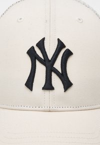 Baseball-Cap aus hellem beigem Stoff mit einem schwarzen gestickten New York Yankees Logo, strukturierter Form und texturiertem Oberflächenfinish.