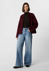 Giacca bordeaux con un fit rilassato e chiusura con zip, abbinata a jeans light blue a vita alta e gamba larga, e stivali neri.