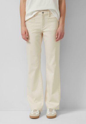 Vorderansicht einer Person, die cremefarbene Hosen mit weitem Bein, weiße Sneakers mit braunen Sohlen und ein weißes, in die Hose gestecktes Oberteil trägt.