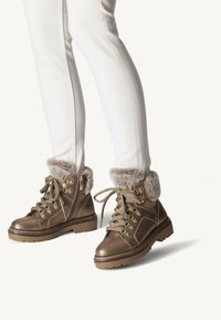 Botas de tobillo de bronce con puños de pelo sintético, cuero texturizado, cordones y cremalleras laterales; diseñadas con suelas resistentes para mayor tracción.
