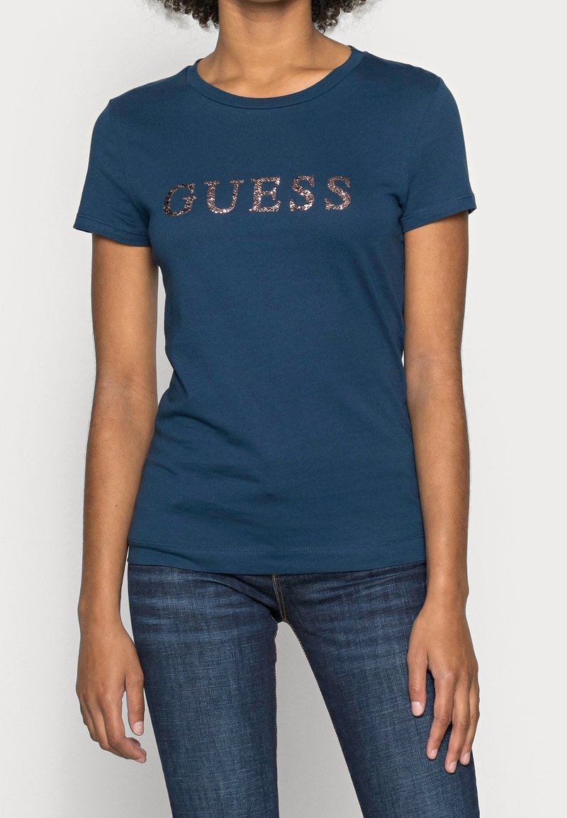 Kvinna iklädd en figurnära marinblå t-shirt med kort ärm med "GUESS" i roséguld paljetter, i kombination med mörkblå denimjeans.