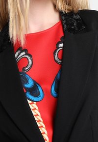 Blazer noir avec un col texturé, porté sur un haut rouge vif orné d'éléments graphiques bleus et d'un motif de chaîne dorée.