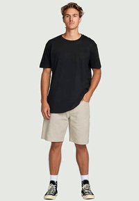 Giovane uomo in piedi, con una semplice t-shirt nera, pantaloni corti beige fino al ginocchio, calze bianche e sneakers nere alte su uno sfondo chiaro.