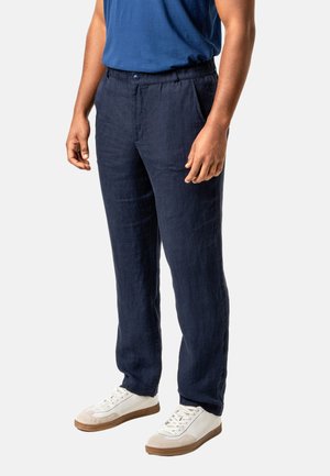 Man wearing navy blue linen pants, blue shirt, and white sneakers standing against a light background.