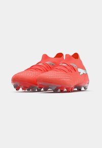 Botas de fútbol de color rojo brillante con una superficie texturizada, que presentan un diseño elegante y un logo plateado como acento. Suela con tacos para tracción.