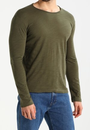 Hombre con camisa ajustada de manga larga verde oliva y vaqueros azules, de pie contra un fondo blanco liso.