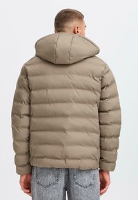 Beige pufferjakke med hætte, der har et quiltet, polstret design. Stoffet fremstår glat med en blød tekstur; minimalistisk stil.