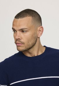 Pull en tricot bleu marine avec un col rond et une bande horizontale blanche sur la poitrine. La texture est lisse et étroitement tissée.