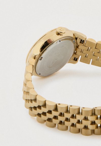 Ρολόι χειρός με χρυσό μεταλλικό μπρασελέ και στρογγυλή πίσω κάσα με κορώνα, εμφανιζόμενο από το πλάι πάνω σε λευκό φόντο.