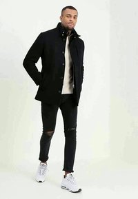 Homem a usar um casaco preto de botões, suéter bege, jeans pretos rasgados e sapatilhas brancas, de pé com as mãos nos bolsos contra um fundo simples.