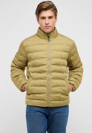 Mann trägt eine hellolivgrüne gesteppte Steppjacke mit den Händen in den Taschen und blaue Jeans vor einem schlichten hellen Hintergrund.