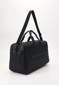 Schwarze Duffel-Tasche mit glatter Textur, doppelten Tragegriffen, verstellbarem Schultergurt und dezentem Logo-Detail an der Seite.