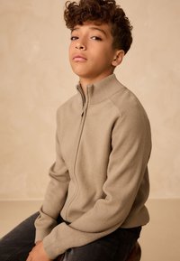 Ragazzo con capelli ricci che indossa un maglione beige con zip e pantaloni scuri, seduto contro uno sfondo beige uniforme, che guarda avanti con sicurezza.