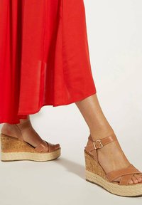 Hakken sandalen in lichtbruin met een kurk en touw textuur basis, voorzien van gekruiste banden en enkelsluitingen. In combinatie met een rode luchtige jurk.