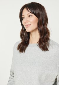 Grijze sweatshirt met een ronde hals en lange mouwen. Onderwerp heeft golvend bruin haar en een neutrale uitdrukking, tegen een lichte achtergrond.