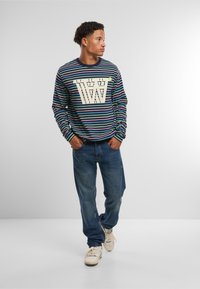 Multikleurige gestreepte lange mouw shirt met een grote witte grafiek, gecombineerd met losse blauwe jeans en witte sneakers. Heeft een geribbelde halslijn.