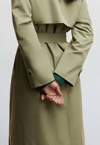 Olivengroene trenchcoat met een ceintuur in de taille en geplooide achterkant; handen gekruist achter de rug; voorzien van manchetdetails met knopen.