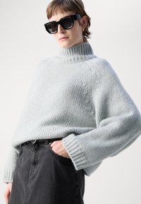 Hladký svetřík světle modré barvy s vysokým límcem, žebrovanými manžetami a spadlými rameny, doplněný černou džínsovou sukni. Na sobě má černé sluneční brýle.