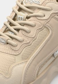 Béžové tenisky s texturovanými vzory, hladkými překryvy a síťovanými detaily. Mají silné šněrování a značkový logo na boku.