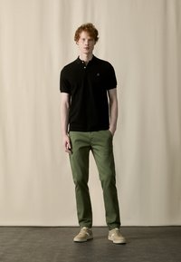 Camiseta de polo negra de manga corta con un pequeño logo bordado, combinada con pantalones verdes y zapatillas beige sobre un suelo gris contra un fondo crema.