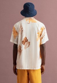 Een man in een shirt met korte mouwen met schelpmotief, mosterdgele broek en een donkerblauwe bucket hat, met zijn rug naar de camera tegen een effen achtergrond.
