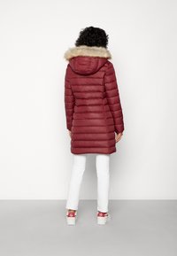 Bordeaux pufferjack met een nepbonten capuchon, verticale gewatteerde textuur, middellange snit, gecombineerd met witte broeken en rode sneakers.