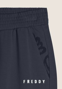 Jogging pantaloni grigio scuro con vita elastica, tasche laterali e un sottile design del logo nero. Presenta una texture del tessuto liscia.