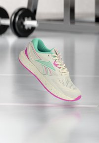 Κρέμ αθλητικό παπούτσι με πράσινες και ροζ πινελιές να αιωρείται πάνω από το δάπεδο ενός γυμναστηρίου, με θολό ράφι βαρών και μπάρα στο φόντο.