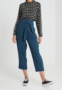 Blusa nera a fantasia con maniche lunghe; pantaloni teal a vita alta con cintura a fiocco. Indossata con sneakers nere e con gambe cropped.