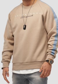 Felpa beige con strisce laterali azzurro chiaro, colletto tondo e logo stampato. Indossata sopra una camicia bianca, abbinata a jeans grigi chiari.