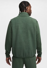 Colete de polar verde com colarinho alto, apresentando um tecido texturizado. Camisa de manga longa verde a condizer visível por baixo do colete.