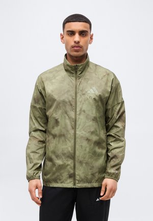 Jonge man draagt een groen camouflagepatroon ritsjack en zwarte broek, staand tegen een effen witte achtergrond.