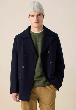 Jeune homme portant un bonnet en maille gris, un manteau croisé bleu marine foncé, un pull vert et un pantalon en velours côtelé couleur fauve sur un fond beige.